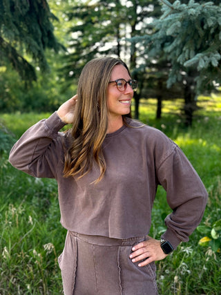 A woman wearing a matching brown long-sleeve crop sweatshirt and sweatpants set, standing outdoors in a grassy area with trees in the background.