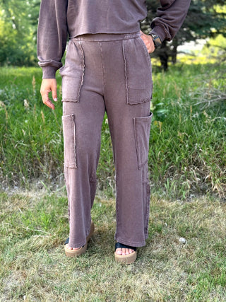 A person standing outdoors on grass wearing loose, wide-leg taupe-brown cargo-style pants with large patch pockets and a matching long-sleeve top.