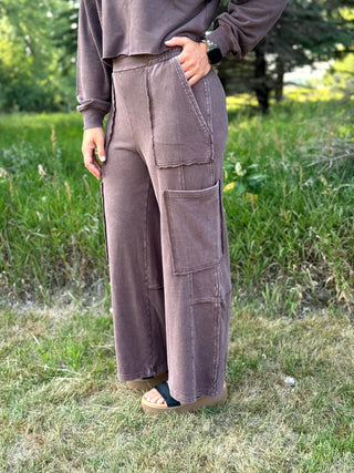 A person standing outdoors on grass wearing loose, wide-leg cargo-style pants in a muted taupe-brown color, paired with a matching long-sleeve top and sandals.