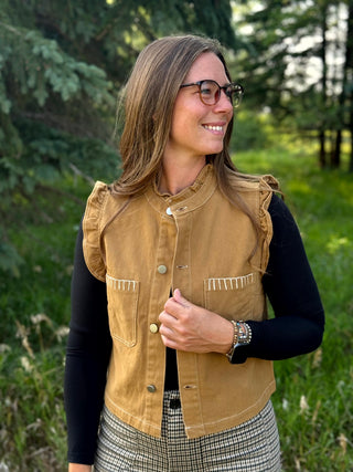 A woman wearing a tan sleeveless vest over a black long-sleeve shirt, standing outdoors in a grassy, wooded area. The vest has a stand-up collar, button front, and two front pockets with decorative stitching.
