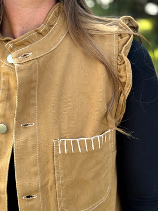 Close-up of a tan canvas vest with white contrast stitching, a stand collar, and a chest pocket, worn by a person with long light brown hair over a black top.