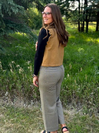 A woman wearing a tan sleeveless ruffle-trimmed top over a black long-sleeve shirt and beige-and-black checkered pants, standing outdoors in a grassy wooded area.