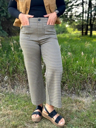 A person standing outdoors in a grassy field wearing high-waisted cropped houndstooth pants in beige and black, a tan sleeveless vest over a black long-sleeve top, and black platform sandals.