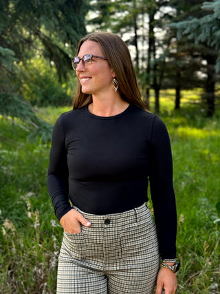 A woman wearing a fitted black long-sleeve top and high-waisted beige-and-black plaid trousers, standing outdoors in a grassy wooded area.