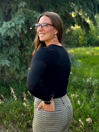 A woman wearing a black long-sleeve top and a black-and-white houndstooth skirt, standing outdoors in a grassy field with trees in the background.