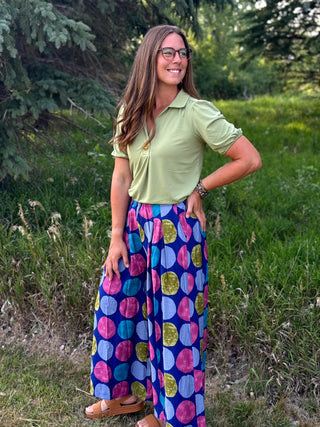 A woman wearing a light green short-sleeve polo shirt and wide-leg blue pants with a colorful pink, purple, blue, and green polka dot pattern, standing outdoors in a grassy field.