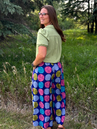A woman standing outdoors in a grassy field, wearing a light sage green short-sleeve blouse and high-waisted, wide-leg pants with a colorful pink, blue, yellow, and green polka dot pattern on a navy base.