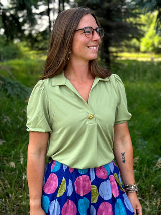 A woman wearing a light green short-sleeve button-up shirt with puff sleeves and a colorful polka-dot skirt, standing outdoors in a grassy area.
