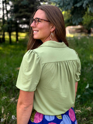 A woman wearing a sage green short-sleeve blouse with puff sleeves and a gathered yoke, shown from the back in an outdoor park setting.