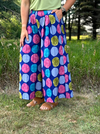 A person standing outdoors in a grassy field wearing wide-leg pleated maxi pants with a colorful abstract dot pattern on a navy blue base, paired with a light green top and brown slide sandals.