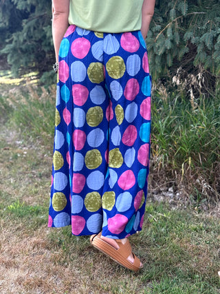 A person outdoors wearing wide-leg pants with a bold multicolor polka dot pattern on a blue base, paired with a light green top and tan sandals.