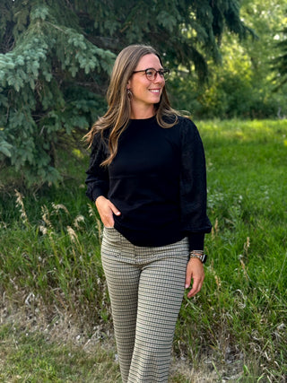 A woman wearing a black long-sleeve top and beige-and-black checkered pants, standing outdoors in a grassy area with trees in the background.