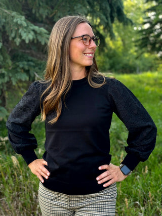 A woman wearing a black crewneck sweater with textured, patterned balloon sleeves, paired with beige and black plaid pants, standing outdoors in a grassy area with trees in the background.