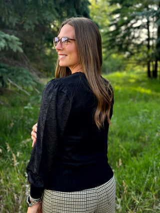 A woman with long brown hair and glasses, wearing a black textured long-sleeve top and a black-and-white houndstooth pant, standing outdoors in a grassy, wooded area.