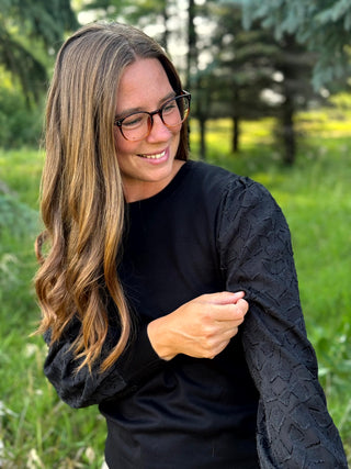 A woman with long brown hair and glasses wearing a black long-sleeve top with textured, patterned sleeves, standing outdoors in a grassy area.