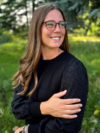 A woman with long brown hair and glasses, wearing a black long-sleeve top with textured lace sleeves, standing outdoors in a grassy area.