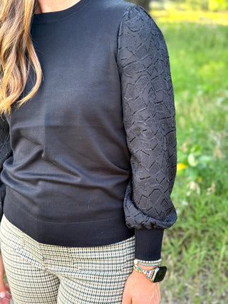 A close-up of a person wearing a black crewneck sweater with textured, patterned long sleeves and black-and-white houndstooth pants, standing outdoors in a grassy area.