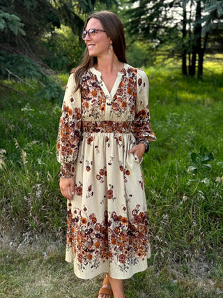 A woman wearing a long-sleeve cream-colored midi dress with a brown and rust floral print, standing outdoors in a grassy wooded area.