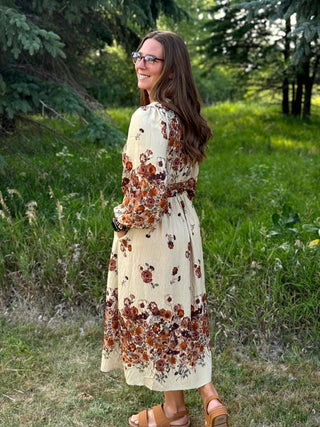 A woman wearing a long-sleeve cream midi dress with a brown and rust floral print, standing outdoors in a grassy wooded area.