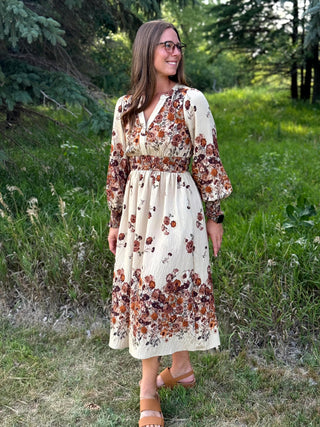 A woman wearing a long-sleeve cream midi dress with a brown and rust floral print, standing outdoors in a grassy wooded area.