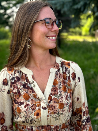 A woman wearing a cream-colored long-sleeve blouse with a brown and rust floral print, styled with tortoiseshell sunglasses and gold hoop earrings, set against a blurred green outdoor background.