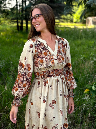 A woman wearing a cream-colored long-sleeve midi dress with a brown and rust floral print, standing outdoors in a grassy park setting.