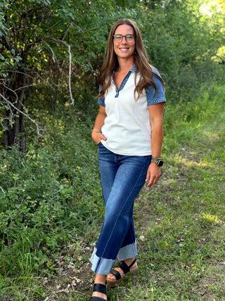 A woman wearing a white and blue color-blocked short-sleeve top, blue jeans, and black sandals stands on a grassy path surrounded by green trees and bushes.
