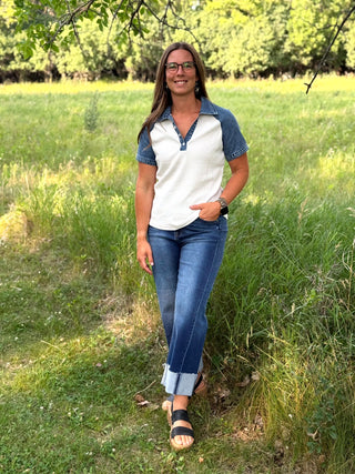A woman wearing a white and blue short-sleeve polo shirt, blue jeans, and black sandals stands in a grassy outdoor field.