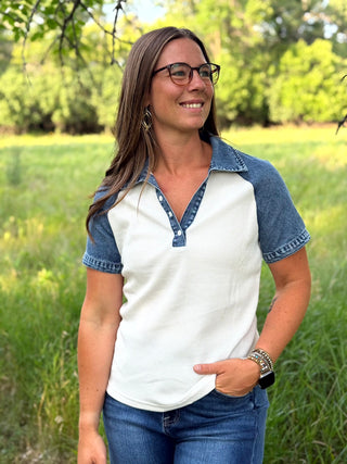 A woman wearing a white and blue raglan henley shirt with denim sleeves and collar trim, standing outdoors in a grassy field.