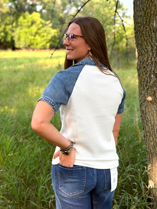 A woman standing outdoors in a grassy field next to a tree, wearing a short-sleeve raglan-style top with a white body and blue denim sleeves and collar, paired with blue jeans.