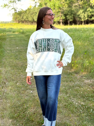 A woman wearing a cream-colored sweatshirt with a black and white patterned 'MINNESOTA' graphic, paired with blue jeans, standing outdoors in a grassy field.