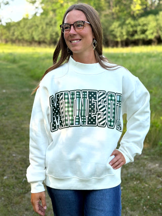 A woman wearing a white crewneck sweatshirt with 'MINNESOTA' printed across the chest in a green plaid and black-and-white striped collegiate font, standing outdoors in a grassy field.