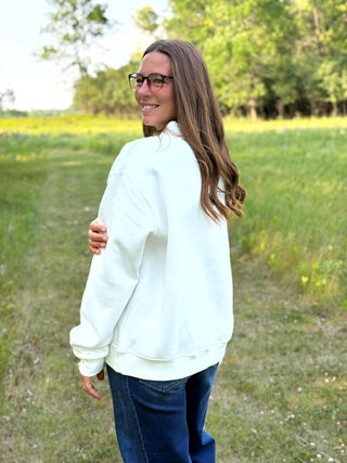 A woman with long brown hair and glasses, wearing a white crewneck sweatshirt and blue jeans, standing in a grassy outdoor field.