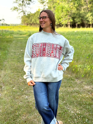 A woman wearing a light gray sweatshirt with a red and white graphic text print that reads 'SOUTHDAKOTA', paired with blue jeans, standing outdoors in a grassy field.