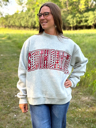 A woman wearing a light heather gray sweatshirt with a red and white patterned 'SOUTH DAKOTA' graphic, standing outdoors in a grassy field.