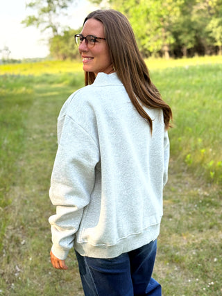 A woman with long brown hair and glasses, wearing a light gray heathered crewneck sweatshirt and dark blue jeans, standing in a grassy outdoor field.