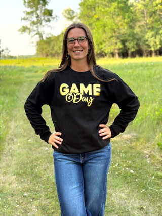 A woman wearing a black crewneck sweatshirt with 'GAME Day' printed in yellow on the front, standing outdoors in a grassy field.