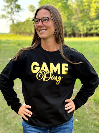 A woman wearing a black long-sleeve sweatshirt with 'GAME Day' printed in gold lettering, standing outdoors in a grassy field with trees in the background.