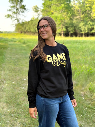 A woman wearing a black crewneck sweatshirt with 'GAME Day' printed in yellow across the chest, standing outdoors in a grassy field.