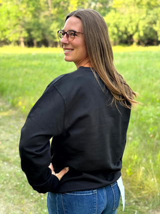 A woman with long brown hair and glasses, wearing a black crewneck sweatshirt and blue jeans, standing outdoors in a grassy field.