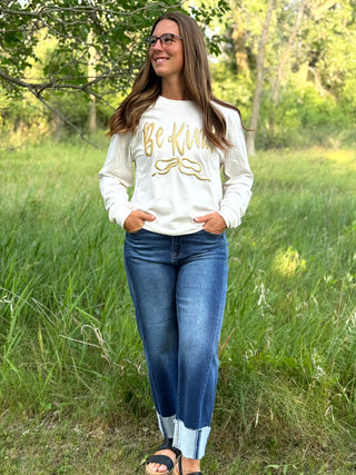 A woman wearing a white long-sleeve crewneck sweatshirt with 'Be Kind' printed in gold script across the chest, paired with blue jeans, standing outdoors in a grassy field with trees in the background.