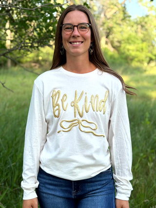 A woman wearing a white long-sleeve crewneck sweatshirt with 'Be Kind' printed in gold script across the chest, paired with blue jeans, standing outdoors in a grassy field with trees in the background.