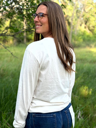 A woman with long brown hair and glasses, wearing a white long-sleeve t-shirt and blue jeans, standing outdoors in a grassy field with trees in the background.