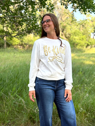 A woman wearing a white long-sleeve crewneck sweatshirt with 'Be Kind' printed in gold script across the chest, paired with blue jeans, standing outdoors in a grassy field with trees in the background.
