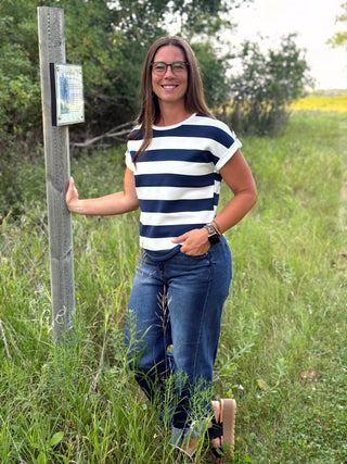 A woman wearing a navy and white striped t-shirt and blue jeans stands outdoors in a grassy field next to a wooden signpost.