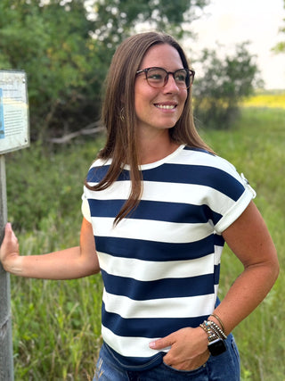 A woman wearing a navy blue and white horizontally striped short-sleeve t-shirt and glasses, standing outdoors in a grassy field next to a signpost.