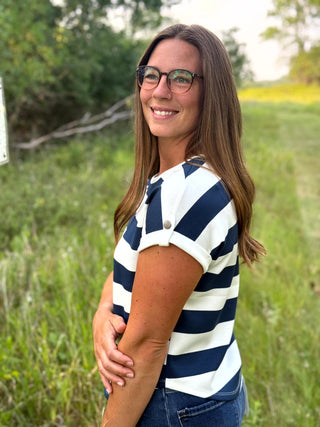 A woman with long brown hair and glasses, wearing a navy and white horizontally striped short-sleeve shirt, standing outdoors in a grassy field.
