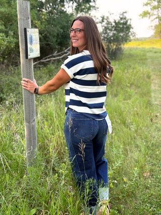 A woman wearing a navy and white striped t-shirt and blue jeans stands outdoors in a grassy field next to a wooden post, looking back over her shoulder.