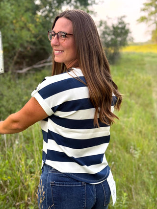 A woman with long brown hair and glasses, wearing a navy and white horizontally striped short-sleeve t-shirt and blue jeans, standing outdoors in a grassy field.