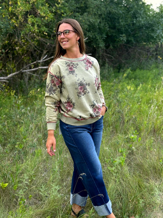 A woman wearing a beige crewneck sweatshirt with a pink and green floral pattern, blue jeans, and glasses, standing in an outdoor grassy field.